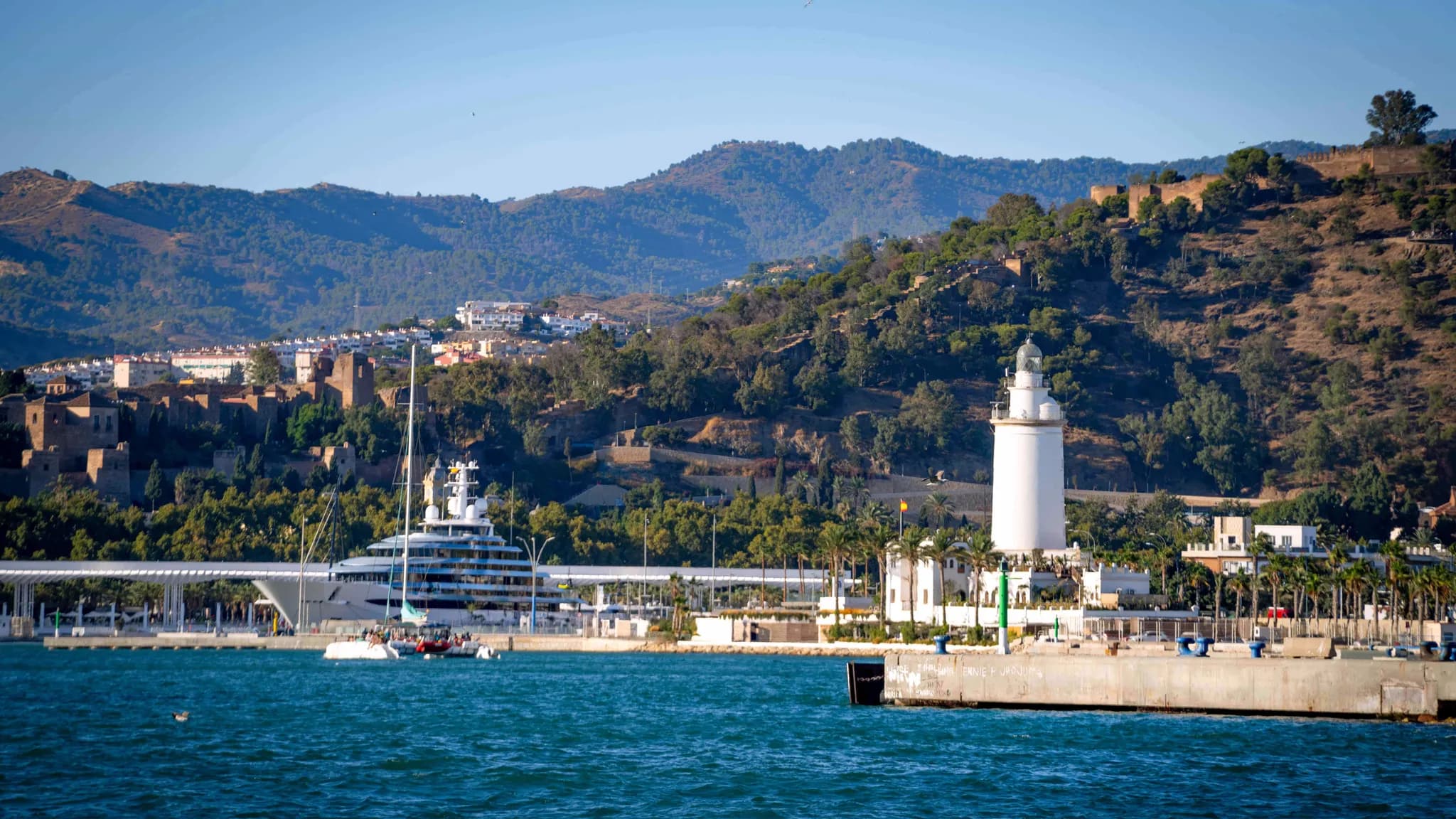 O porto de Málaga com o farol La Farola, um superiate no cais e a Alcazaba na encosta arborizada