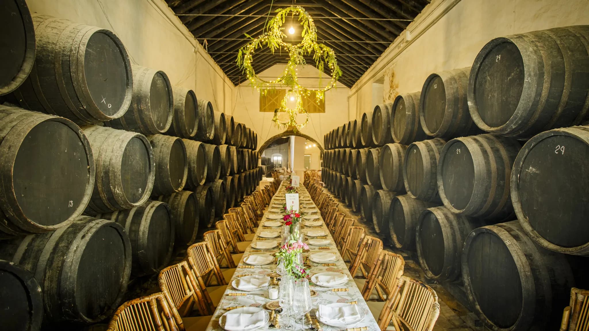 Uma longa mesa de banquete posta entre filas de barris numa adega, sob um lustre de flores