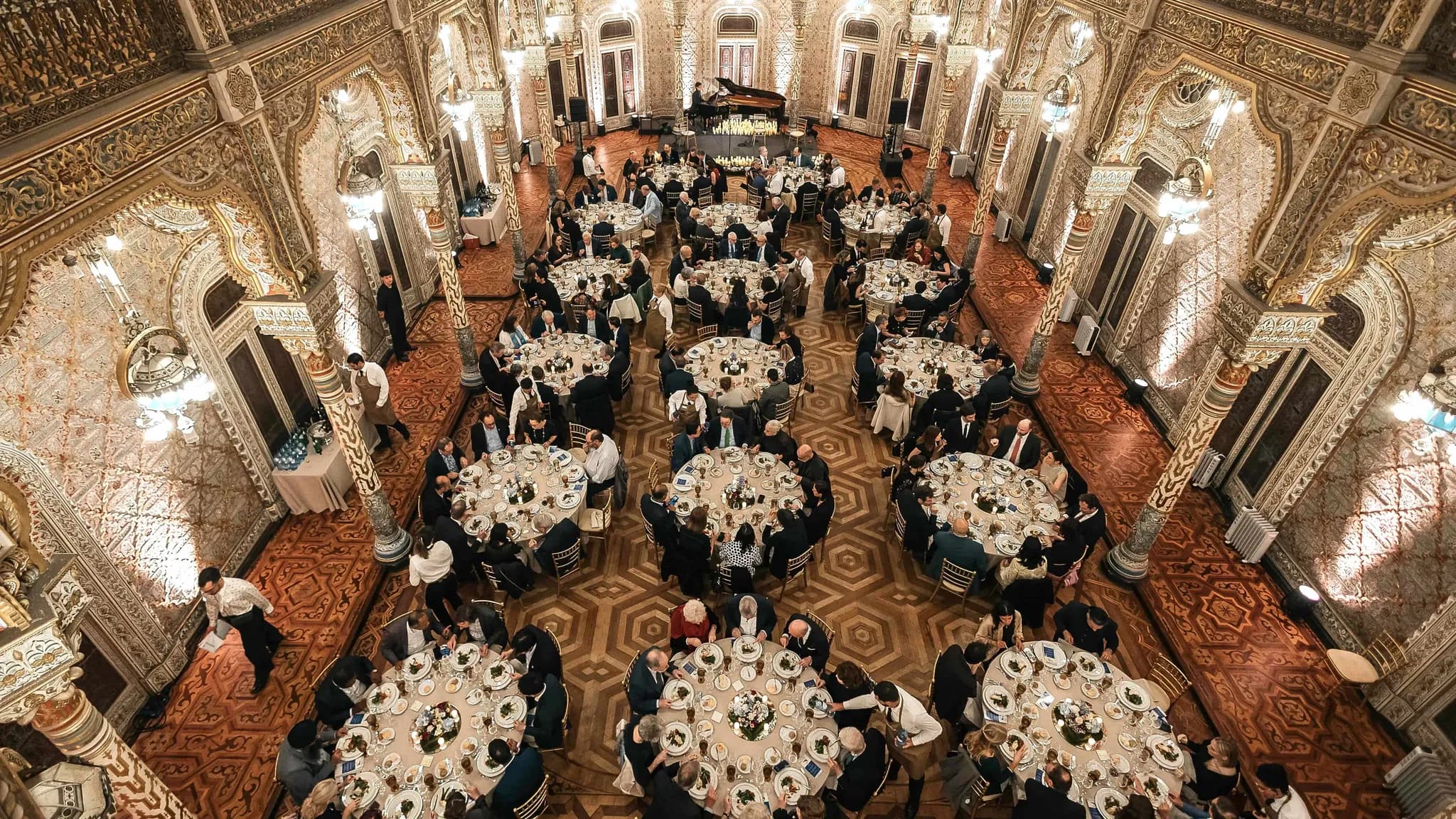 Jantar de gala num salão ornamentado de estilo árabe, com convidados em mesas redondas sob candeeiros de cristal