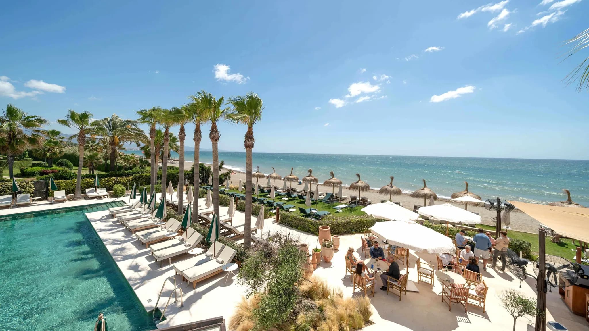 Piscina de um hotel junto à praia, rodeada de palmeiras e espreguiçadeiras, com convidados no terraço à beira-mar